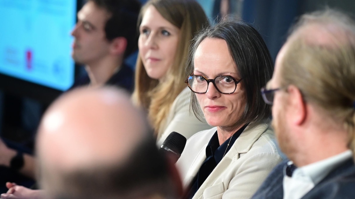 Delia Strunz mit einem Mikrofon in der Hand und weiteren Speakern neben sich. Sie hat kinnlange Haare und trägt eine Brille.