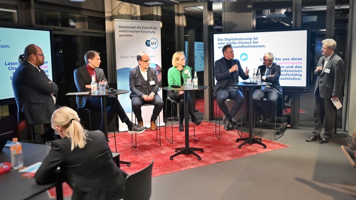 Sechs Personen sitzen nebeneinander auf hohen Stühlen; ein Mann unter ihnen hält ein Mikrofon und spricht, während die anderen Anwesenden zuhören. Hinter ihnen befinden sich zwei Bildschirme und ein TMF-Roll-up.