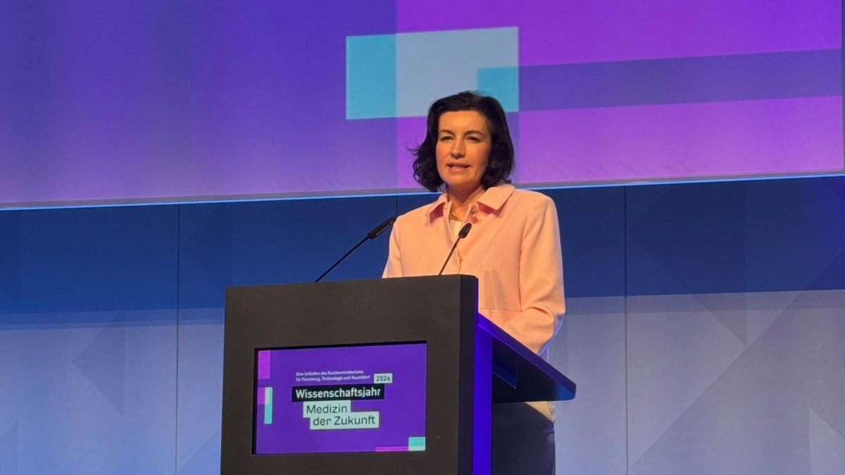 Eine Frau steht an einem Rednerpult und hält eine Rede auf einer Bühne. Sie trägt ein hellrosa Sakko, vor ihr sind zwei Mikrofone. Im Hintergrund ist eine farbige Projektion zu sehen; auf dem Pult steht der Schriftzug ‚Wissenschaftsjahr 2026 – Medizin der Zukunft‘.