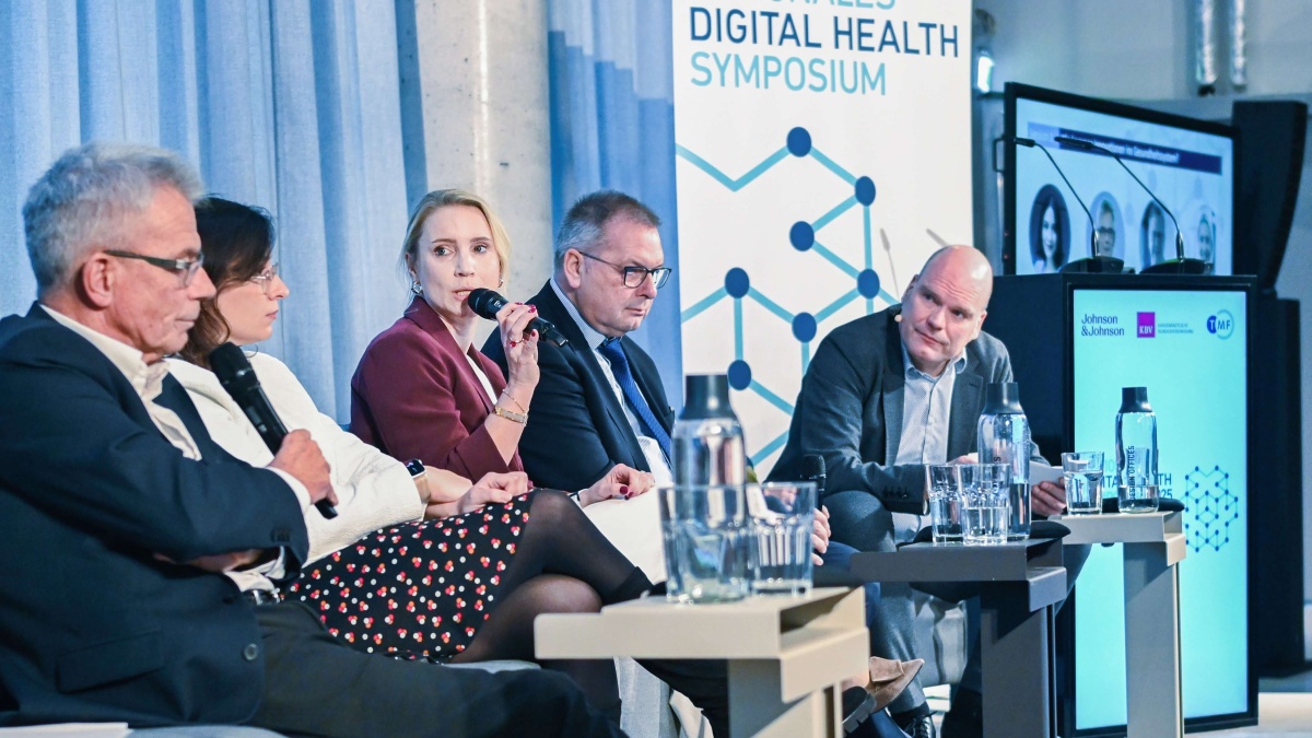 Podiumsdiskussion beim Nationalen Digital Health Symposium mit fünf Personen auf der Bühne. Eine Person spricht in ein Mikrofon, im Hintergrund sind das Veranstaltungsbanner und ein Bildschirm mit Fotos der Speaker zu sehen.