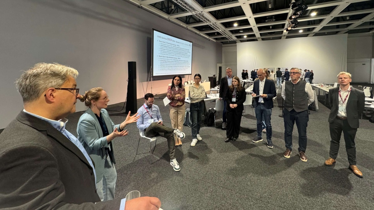 Kleine Gruppe von Teilnehmenden diskutiert in einem Konferenzraum; eine Frau spricht gestikulierend, während andere zuhören, im Hintergrund sind Tische und eine Präsentationsfolie zu sehen.