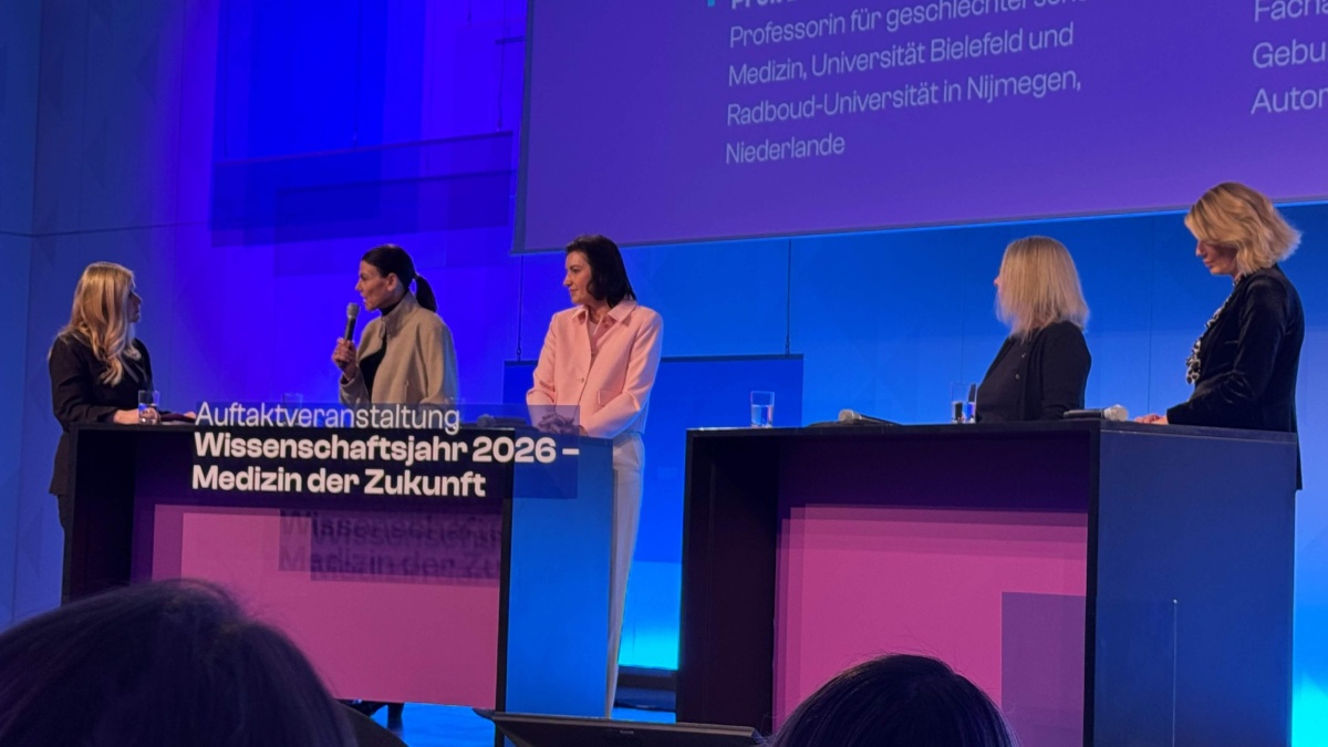 Fünf Frauen stehen auf einer Bühne hinter zwei Stehpulten und nehmen an einer Podiumsdiskussion teil. Eine der Frauen spricht in ein Handmikrofon, die anderen hören zu. Die Bühne ist blau und violett beleuchtet. Auf dem linken Pult steht der Schriftzug ‚Auftaktveranstaltung Wissenschaftsjahr 2026 – Medizin der Zukunft‘, im Hintergrund ist eine Präsentationsfolie zu sehen.