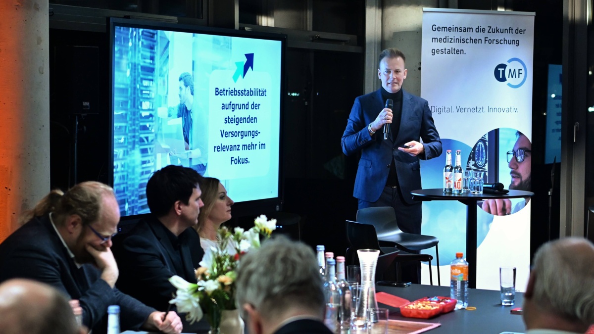 Eine Mann steht mit einem Mikrofon vor einem Publikum. Links von ihm ist ein Bildschirm mit Präsentation und rechts ein TMF-Roll-Up. Mehrere Personen sitzen vor ihm, hören zu und machen sich Notizen; auf dem Tisch stehen Getränke und Blumenarrangements.