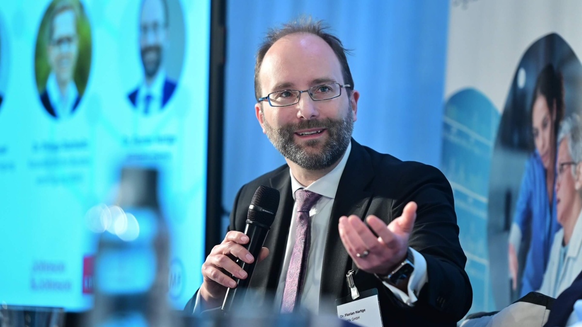 Dr. Florian Hartge mit dunklem Anzug und Brille. Er hält ein Mikrofon und gestikuliert mit seiner freien Hand. Im Hintergrund ist ein unscharfer Bildschirm mit einer PowerPoint-Präsentation zu erkennen.