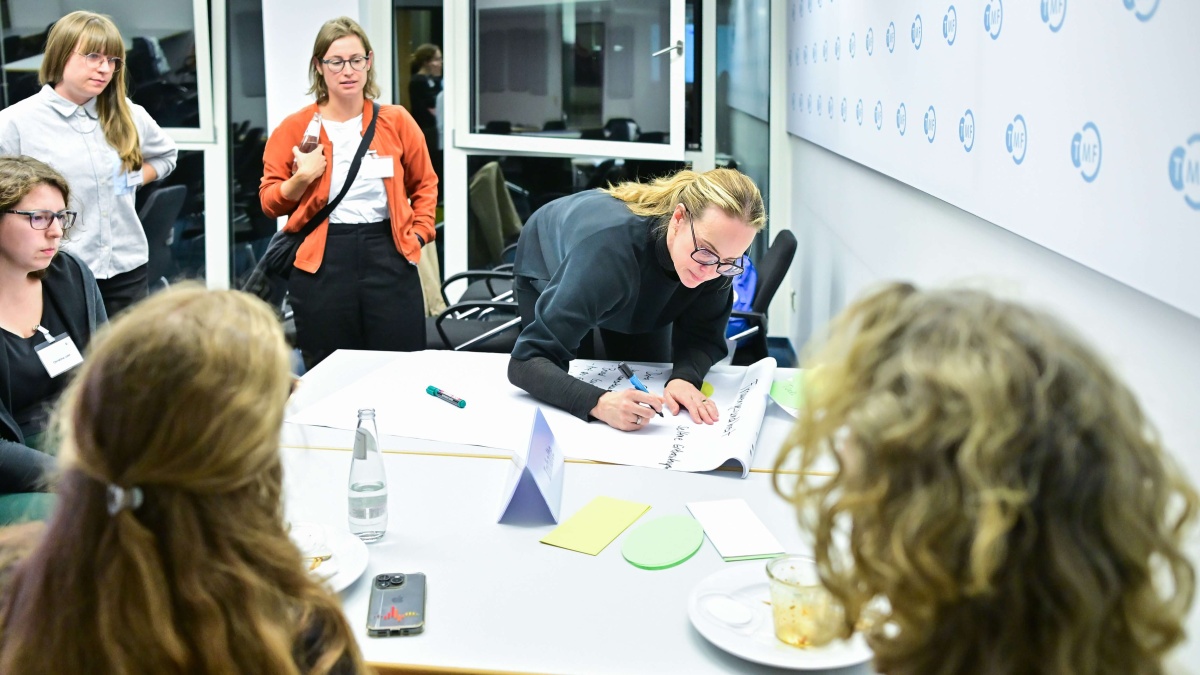 Teilnehmende arbeiten während des Kick-offs gemeinsam an einem Workshop-Tisch. Eine Person notiert Ideen auf einem großen Moderationspapier, während weitere Teilnehmende die Ergebnisse diskutieren und beobachten.
