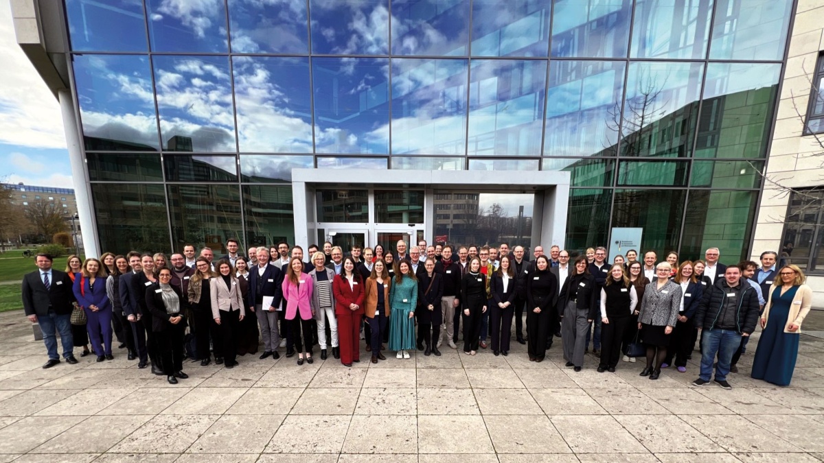 Großes Gruppenfoto vor dem modernen Glasgebäude des BfArM in Bonn. Die Personen stehen in mehreren Reihen vor dem Haupteingang, lächeln in die Kamera und tragen überwiegend Business-Kleidung. Die Glasfassade spiegelt den blauen Himmel mit Wolken.