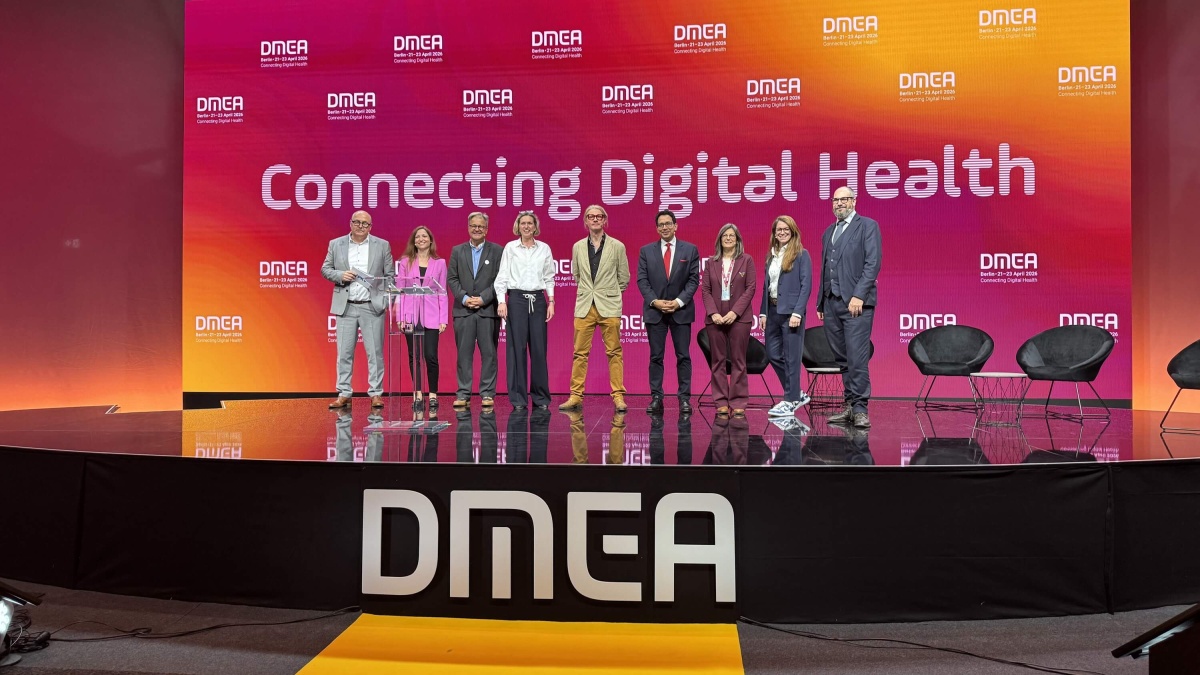 Gruppe von neun Personen steht auf einer Bühne vor einem großen Hintergrund mit der Aufschrift „Connecting Digital Health“ und „DMEA“; sie posieren gemeinsam für ein Foto.