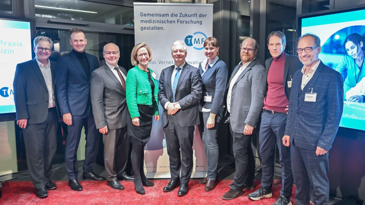 Neun Personen stehen nebeneinander und lächeln in die Kamera. Neben und hinter ihnen befinden sich Bildschirme und ein TMF-Roll-up.