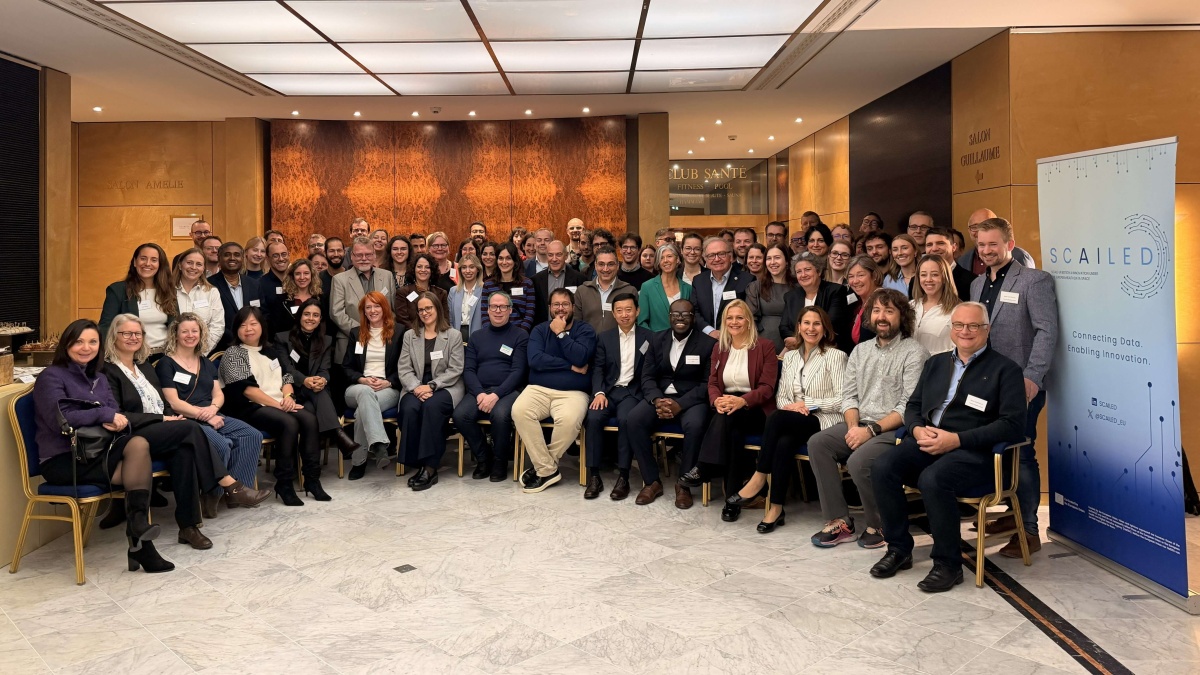 Gruppenfoto der Teilnehmenden des SCAILED-Kick-offs. Die Gruppe ist in einem Hotel- oder Konferenzbereich versammelt und blickt in die Kamera. Rechts im Bild steht ein SCAILED-Roll-up-Banner mit dem Slogan „Connecting Data. Enabling Innovation.“.
