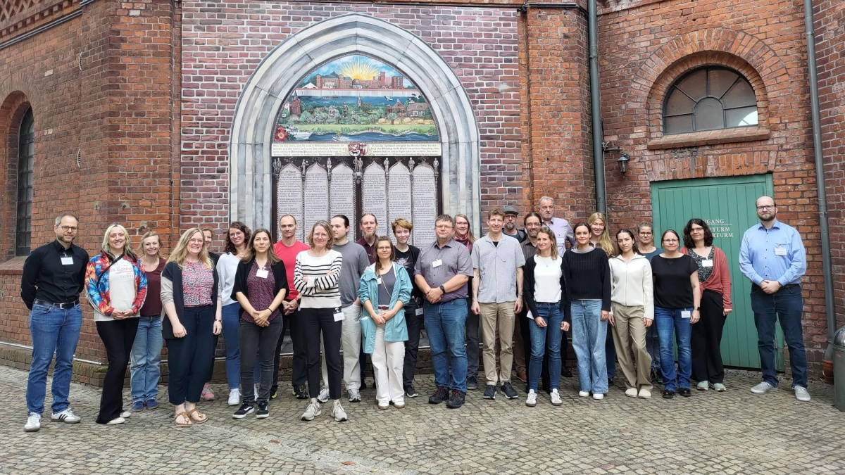 Gruppenfoto von rund 30 Personen, die vor einer historischen Backsteinfassade mit einem großen gotischen Bogenportal stehen. Im Bogen befindet sich eine kunstvoll gestaltete Tafel mit einem farbigen Mosaik und Inschriften. Die Gruppe blickt überwiegend in die Kamera und ist in mehreren Reihen angeordnet. Die Teilnehmenden tragen Alltagskleidung, einige haben Namensschilder um den Hals. Rechts ist eine grüne Tür in die Backsteinfassade eingelassen, im Vordergrund befindet sich ein gepflasterter Platz.