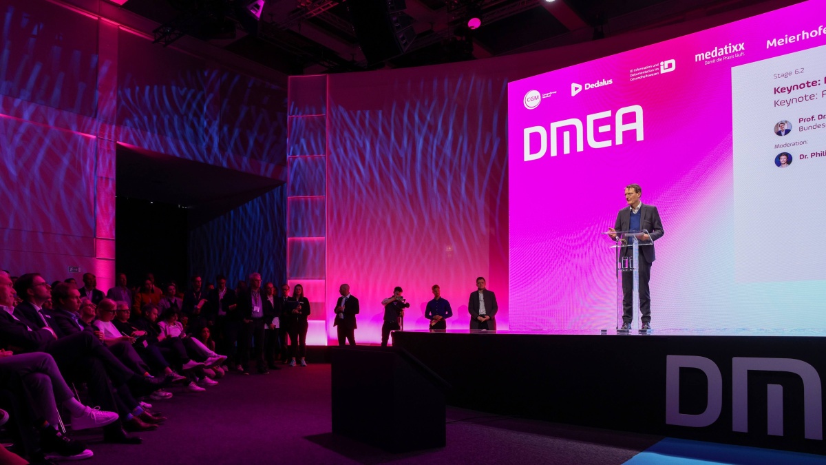 Ein Redner steht auf einer Bühne mit großem pink beleuchtetem Hintergrund und dem Schriftzug „DMEA“. Er spricht an einem transparenten Rednerpult vor einem sitzenden Publikum auf der linken Seite.