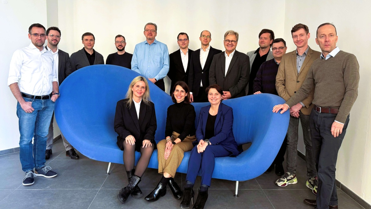 Mehrere Personen stehen hinter einer modernen, blauen Couch, auf der drei Personen sitzen. Sie lächeln für das Gruppenfoto in die Kamera.