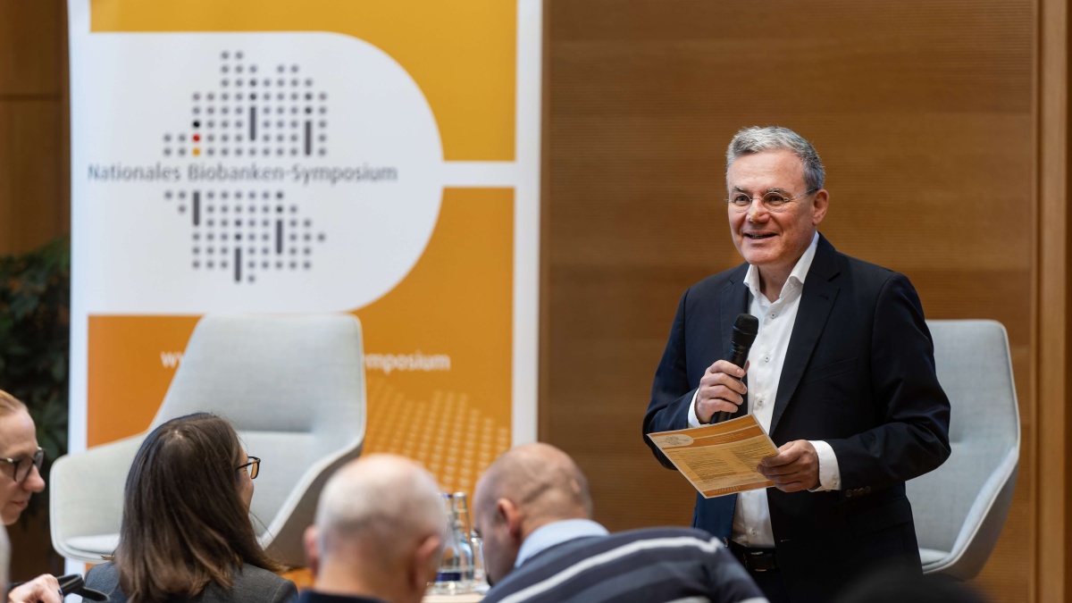 Ein Mann im dunklen Anzug steht mit Mikrofon und Unterlagen vor einem Publikum und spricht auf dem Nationalen Biobanken-Symposium. Im Hintergrund sind zwei helle Sessel sowie ein orange-weißes Veranstaltungsbanner mit dem Logo des Symposiums zu sehen.