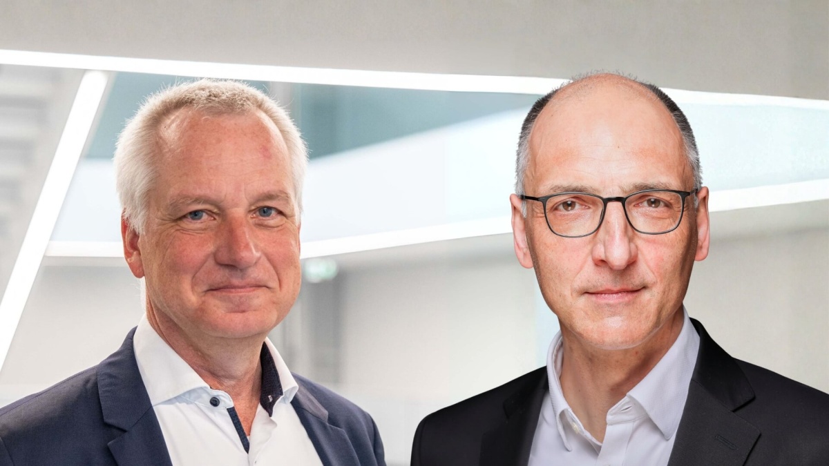 Zwei Männer in Anzügen stehen nebeneinander vor einem modernen, hellen Hintergrund mit diagonalen Lichtlinien. Der Mann links hat graues Haar und trägt ein dunkelblaues Sakko mit weißem Hemd. Der Mann rechts ist glatzköpfig, trägt eine Brille und ein schwarzes Sakko mit weißem Hemd. Beide blicken freundlich in die Kamera.