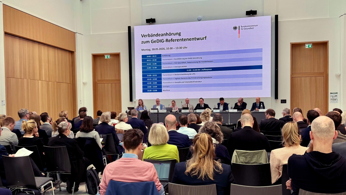 Großer Konferenzsaal mit zahlreichen sitzenden Teilnehmenden bei einer Anhörung des Bundesgesundheitsministeriums. Vorne sitzt ein Podium mit mehreren Sprecherinnen und Sprechern. Auf einer großen Leinwand steht „Verbändeanhörung zum GeDIG-Referentenentwurf“ mit einem Zeitplan für die Veranstaltung am 18.05.2026.