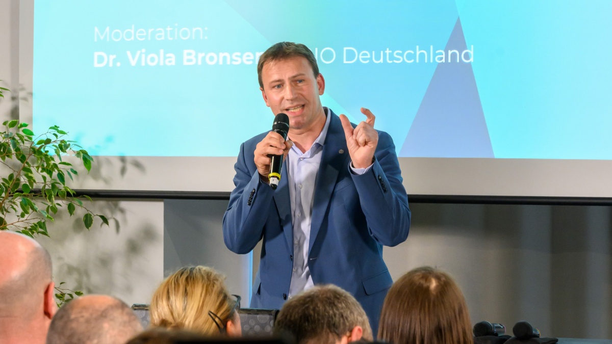 Ein Mann mit Anzug und Mikrofon spricht zu einem Publikum.