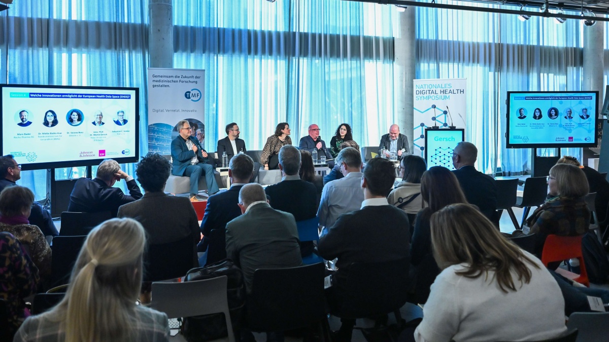 Blick auf eine Podiumsdiskussion beim Nationalen Digital Health Symposium. Mehrere Personen sitzen auf der Bühne und diskutieren vor einem voll besetzten Publikum. Thema der Session sind Innovationen durch den European Health Data Space (EHDS).