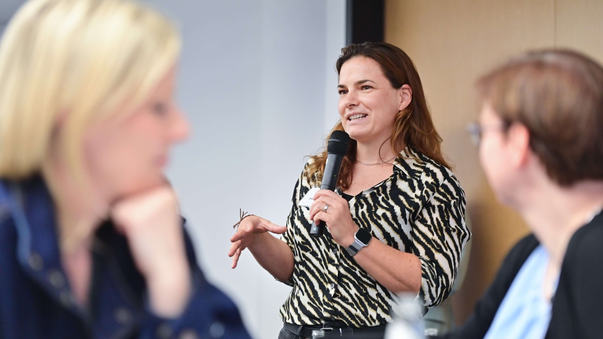 Dr. Ines Reinecke spricht während des Kick-offs in ein Handmikrofon und gestikuliert mit der freien Hand. Im Vordergrund sind mehrere Teilnehmende unscharf zu sehen.