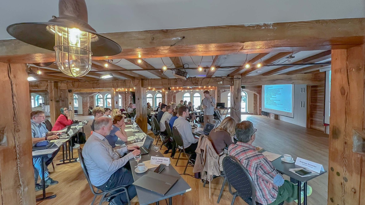 TMF-School 2025 Ein großer Veranstaltungsraum, in dem die Teilnehmenden der TMF-School 2025 an mehreren Tischen sitzen, während ein Mann vor einer PowerPoint zu ihnen spricht.