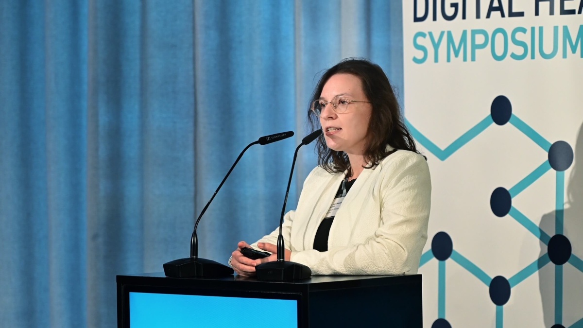 Dr. Jacqueline Lammert, welche in ein Mikrofon spricht. Sie hat schulterlanges Haar und trägt eine Brille. Im Hintergrund ist ein unscharfer Bildschirm mit einer PowerPoint-Präsentation zu erkennen.