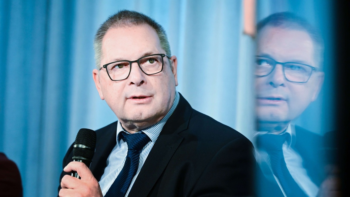 BfArM-Präsident Prof. Karl Broich mit Anzug und Brille. Er hält ein Mikrofon und blickt leicht zur Seite. Rechts im Bild spiegelt sich sein Gesicht an einer Glasfläche. Der Hintergrund besteht aus blauen Vorhängen.