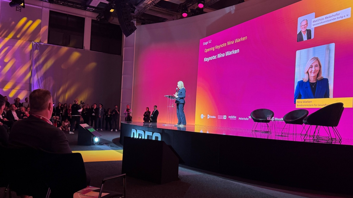 Eine Frau spricht bei einer Keynote auf einer Bühne vor Publikum; hinter ihr eine große, pink-orange Präsentationswand mit ihrem Porträt und Namen, mehrere Stühle stehen rechts auf der Bühne.