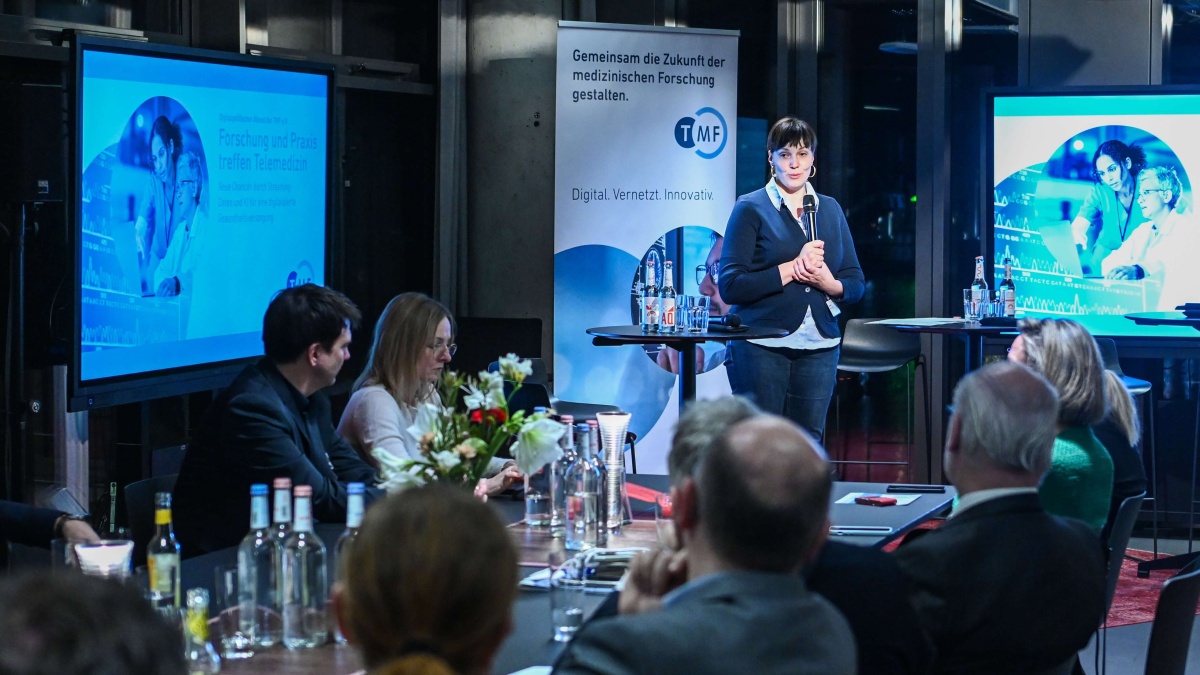 Eine Frau steht mit einem Mikrofon vor einem Publikum. Hinter ihr stehen zwei Bildschirme und ein TMF-Roll-Up. Mehrere Personen sitzen an Tischen, hören zu und machen sich Notizen; auf den Tischen stehen Getränke und Blumenarrangements.