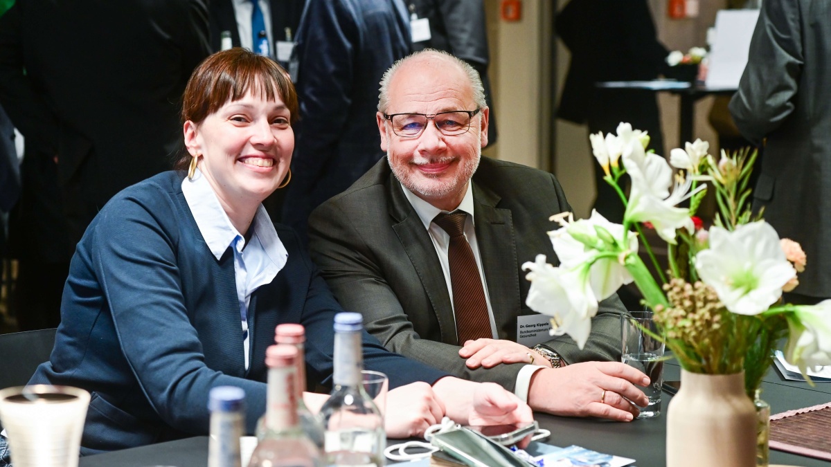 Eine Frau und ein Mann sitzen nebeneinander an einem Tisch und lächeln herzlich in die Kamera. Vor ihnen stehen Getränkeflaschen, Gläser und ein Blumenarrangement in einer Vase. Im Hintergrund sind weitere Personen in Gesprächssituationen zu sehen.