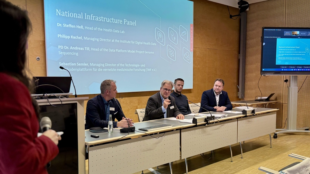 Vier Männer sitzen an einem langen Tisch mit Mikrofonen. Hinter ihnen ist eine PowerPoint mit dem Titel "National Infrastructure Panel" zu sehen.