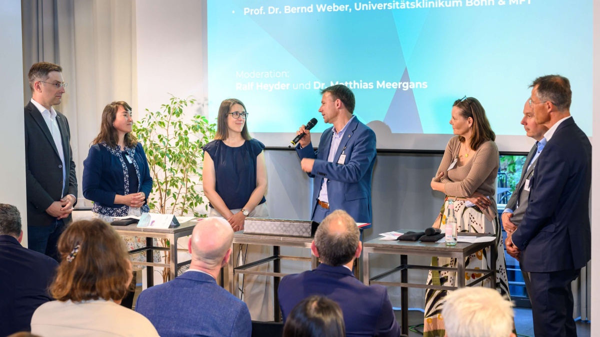 Sechs Personen stehen vor Publikum im Rahmen einer Podiumsdiskussion. Ein Mann in der Mitte spricht in ein Mikrofon. Im Hintergrund wird eine PowerPoint ausgestrahlt.