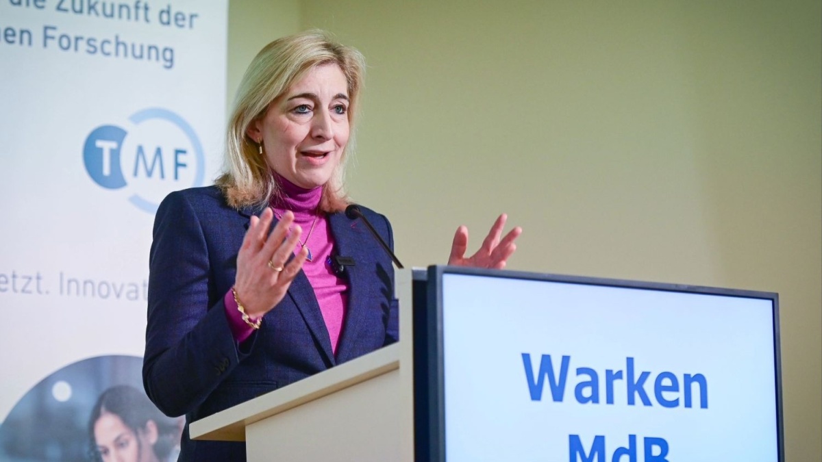 Eine Frau steht an einem Rednerpult und spricht mit erhobenen Händen zu einem Publikum. Sie trägt ein Sakko über einem farbigen Rollkragenpullover. Auf dem Pult ist ein Bildschirm mit dem Text ‚Warken MdB‘ zu sehen. Im Hintergrund befindet sich ein Banner mit dem Logo ‚TMF‘.