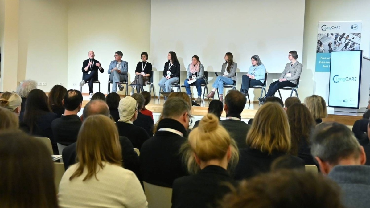 Acht Personen sitzen nebeneinander auf einer Bühne. Vor ihnen befindet sich das Publikum. Rechts stehen ein Roll-up und ein Bildschirm mit dem LongCARE-Logo.