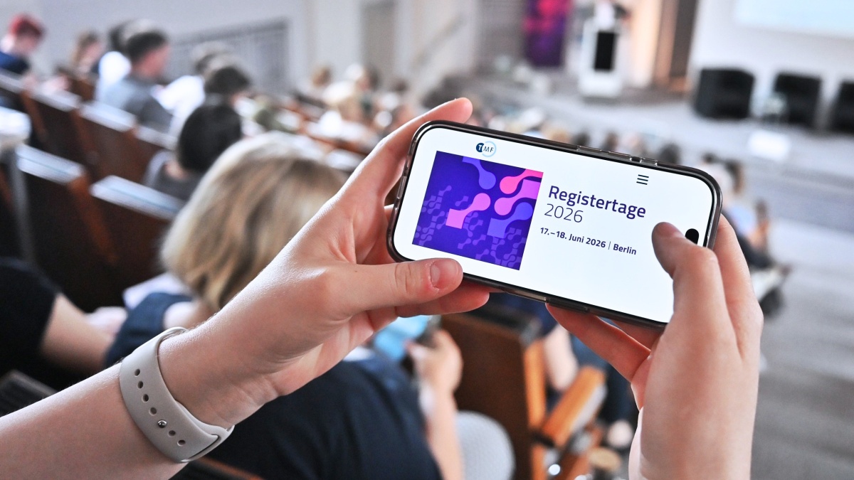 Zwei Hände halten ein Handy, auf dem das Banner der Registertage 2026 zu sehen ist. Im Hintergrund ist der Veranstaltungssaal mit mehreren Teilnehmenden zu erkennen.