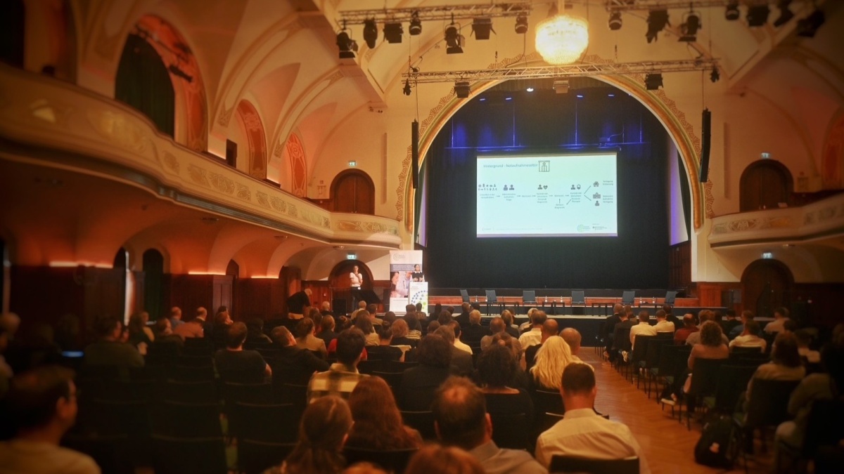 Das Volkshaus Jena von innen. Ein großer Saal mit Publikum, welches einem Vortrag mit PowerPoint zuhört.