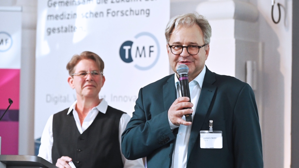 Ein Mann im Anzug spricht in ein Handmikrofon, während eine weitere Person neben ihm steht. Im Hintergrund sind Roll-up-Banner der TMF e.V. zu sehen.