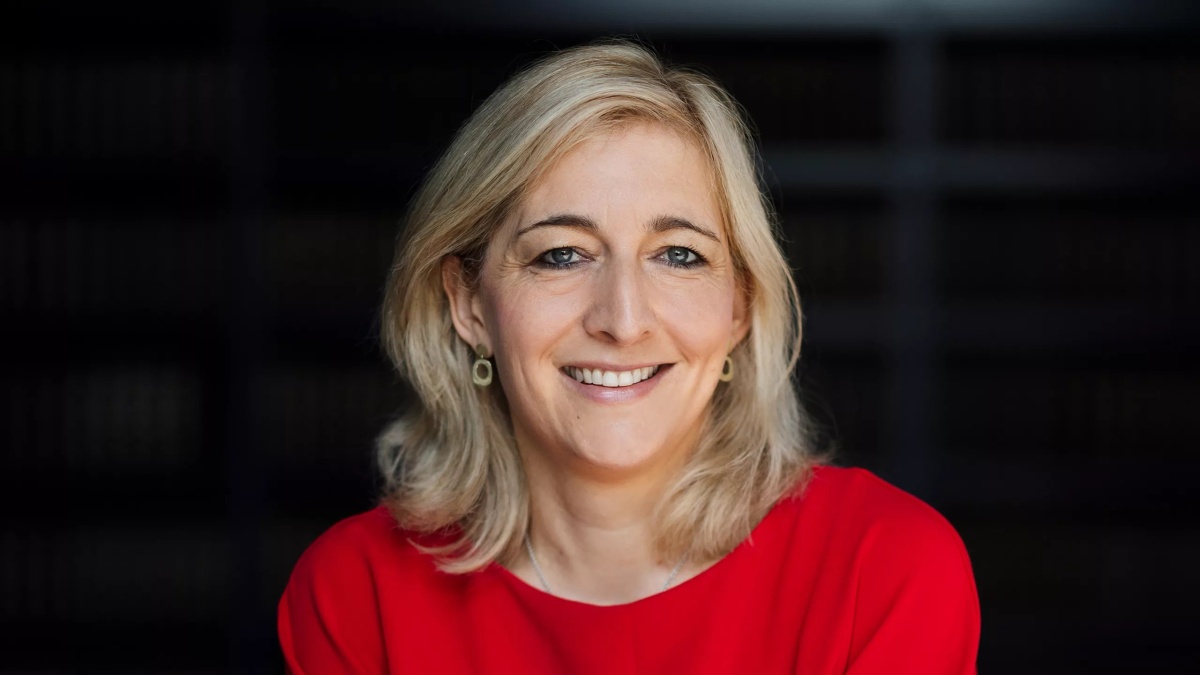 Porträt einer lächelnden Frau mit schulterlangem blondem Haar, die ein rotes Oberteil trägt. Der Hintergrund ist dunkel.