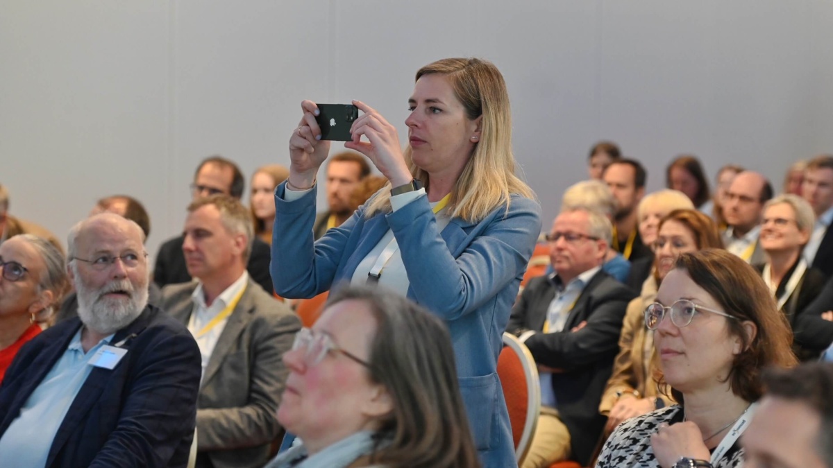 Publikum Eine stehende Frau im Publikum fotografiert mit einem Handy in beiden Händen.
