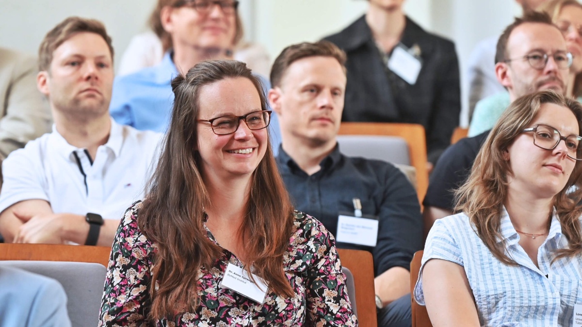 Teilnehmende der Registertage sitzen in einem Hörsaal und verfolgen aufmerksam einen Vortrag. Im Vordergrund lächelt eine Frau mit Brille, während weitere Personen konzentriert zuhören.