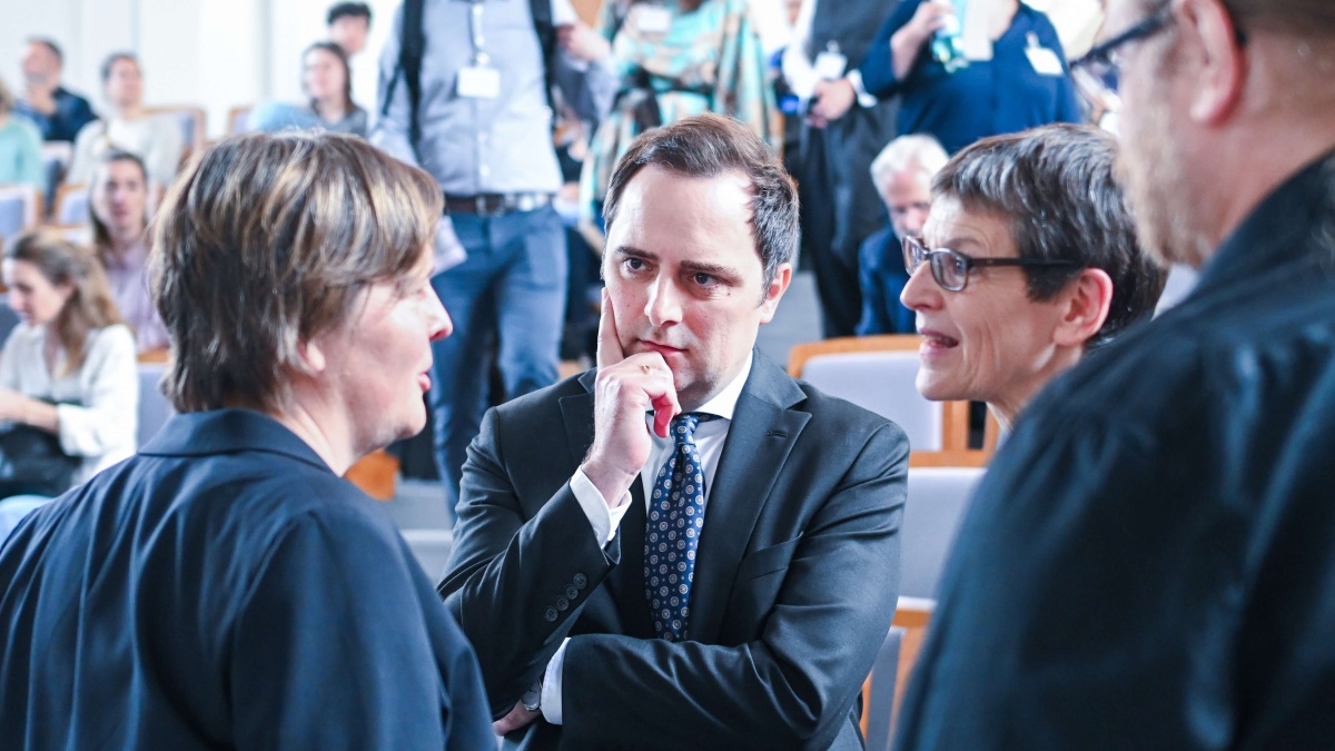 Mehrere Personen in formeller Geschäftskleidung stehen in einem Hörsaal zusammen und führen ein Gespräch. Im Vordergrund diskutieren zwei Frauen mit einem Mann im Anzug, während im Hintergrund weitere Teilnehmende einer Veranstaltung zu sehen sind.