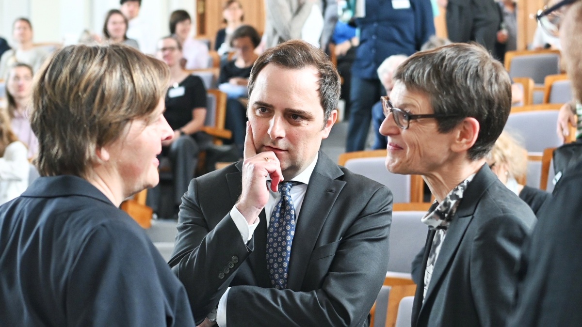 Mehrere Personen in formeller Geschäftskleidung stehen in einem Hörsaal zusammen und führen ein Gespräch. Im Vordergrund diskutieren zwei Frauen mit einem Mann im Anzug, während im Hintergrund weitere Teilnehmende einer Veranstaltung zu sehen sind.