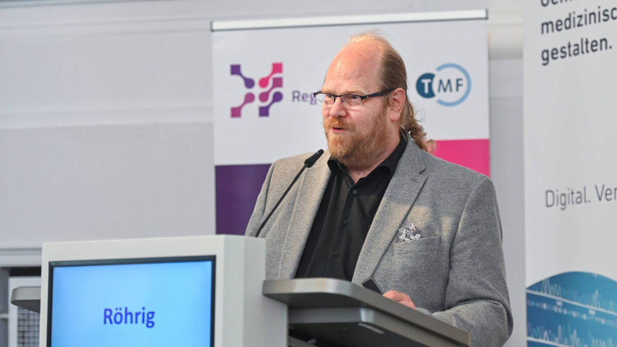 Ein Mann mit grauem Anzug und schwarzem Hemd steht an einem Rednerpult mit der Aufschrift "Röhrig". Hinter ihm sind Roll-up-Banner mit den Logos der Registertage und TMF e.V. zu sehen.