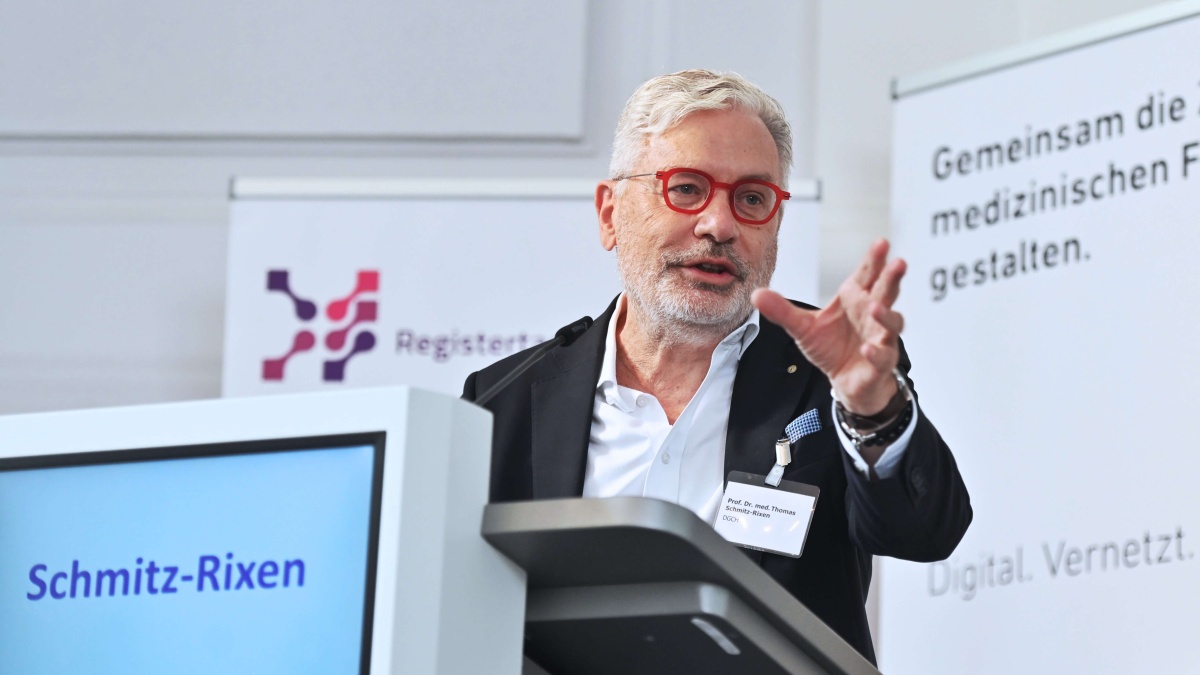 Ein Mann mit Anzug und roter Brille steht an einem Rednerpult mit der Aufschrift "Schmitz-Rixen". Im Hintergrund sich Roll-up-Banner der Registertage und TMF e.V. zu sehen.