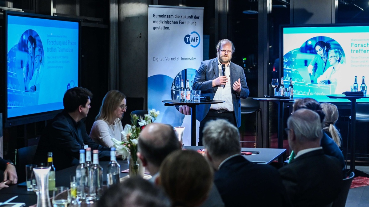 Eine Mann steht mit einem Mikrofon vor einem Publikum. Hinter ihm stehen zwei Bildschirme und ein TMF-Roll-Up. Mehrere Personen sitzen an Tischen, hören zu und machen sich Notizen; auf den Tischen stehen Getränke und Blumenarrangements.