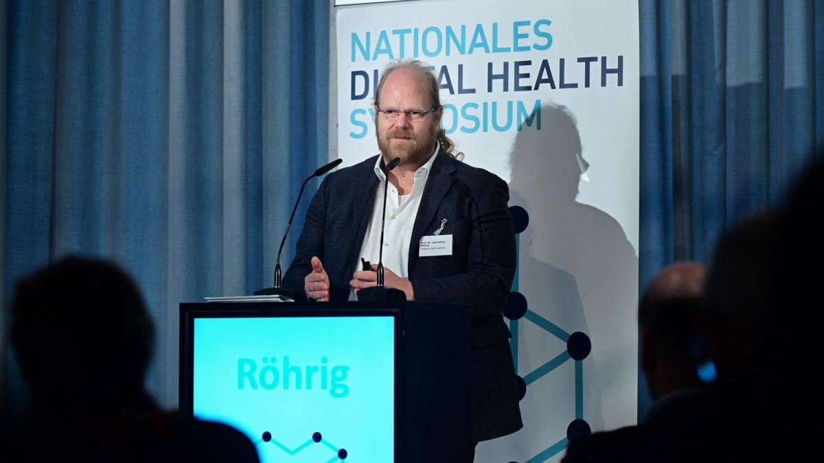 Prof. Dr. Rainer Röhrig vor einem Rednerpult. Im Hintergrund ist ein Banner mit dem Text ‚Nationales Digital Health Symposium‘ zu sehen.