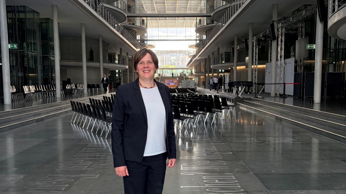 Eine lächelnde Frau im Business-Look steht in einem modernen Gebäude mit zahlreichen Sitzreihen hinter sich, die für eine Veranstaltung platziert wurden.