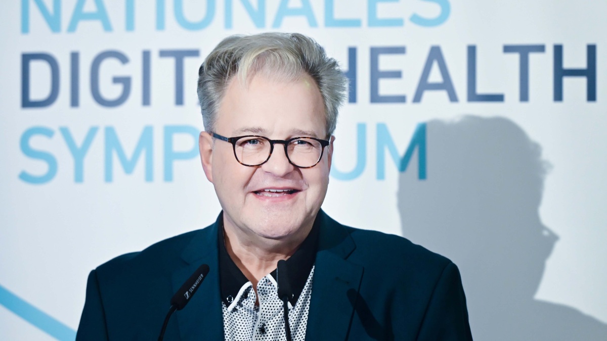 Sebastian C. Semler mit Anzug und Brille. Er spricht in zwei Mikrofone. Im Hintergrund steht der Text ‚Nationales Digital Health Symposium‘.