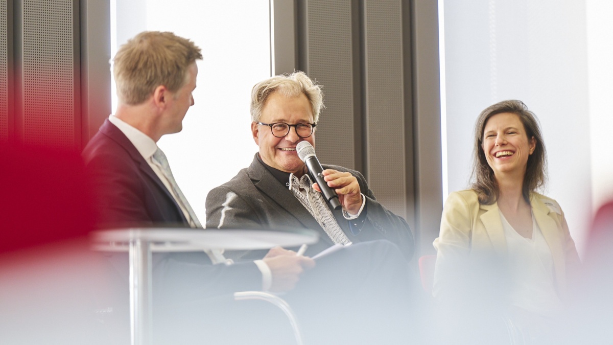 Drei Personen sitzen auf einer Bühne bei einer Podiumsdiskussion. Eine Person in der Mitte spricht in ein Handmikrofon, während die beiden anderen Personen zuhören. Der Raum ist hell und von großen Fenstern geprägt.