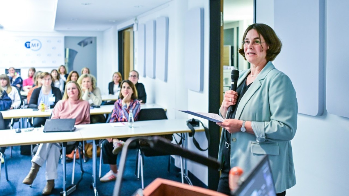 Dr. Stefanie Märschenz steht mit einem Handmikrofon und Unterlagen beim Kick-off vor den Teilnehmenden. Die Zuhörenden sitzen an Tischen und hören zu. Im Hintergrund ist das TMF-Logo an der Wand zu erkennen.