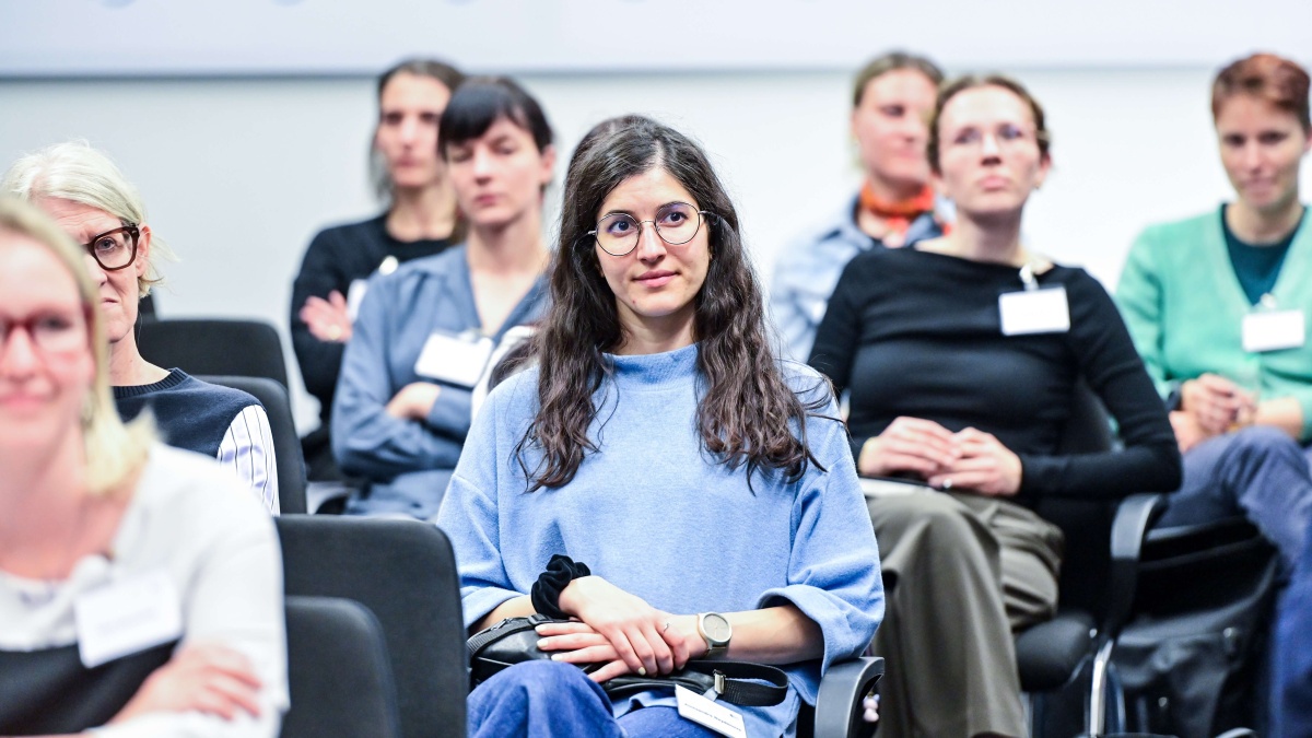 Mehrere Teilnehmende sitzen in einem Veranstaltungsraum und hören einem Vortrag zu. Im Mittelpunkt des Bildes sitzt eine Person mit Brille und blauem Oberteil in einer Zuschauerreihe, während weitere Teilnehmende im Hintergrund zu sehen sind.