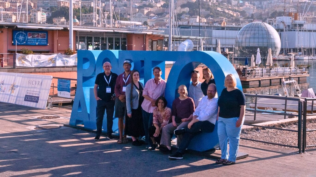Zehn Personen stehen vor dem großen Text "Porto Antico" am Hafen in Genua und lächeln in die Kamera.